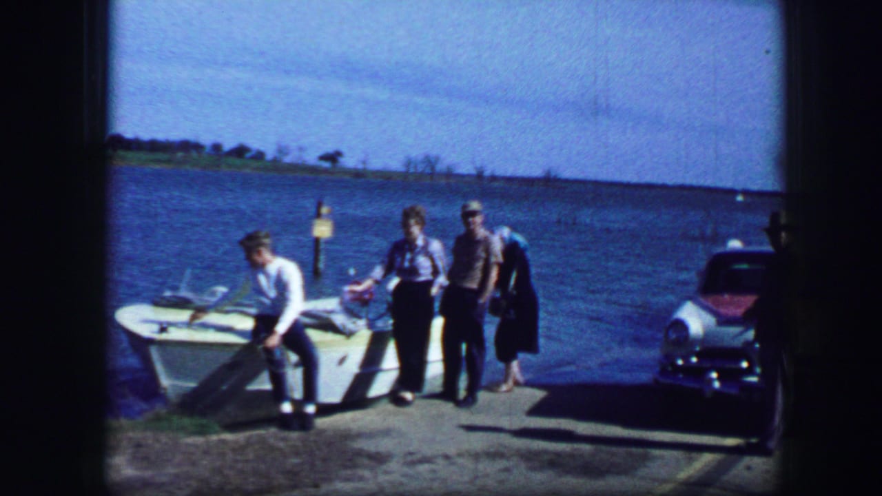 Three adults stand near a small boat at a lakeside dock while one bends toward the bow.