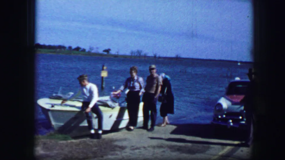 1964 Fort Worth-area home-movie frame showing three adults beside a small boat on a dock with lake water and a parked car nearby.