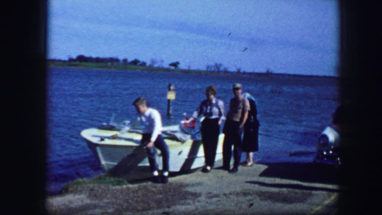 A person bends over the boat while two others stand beside the dock near the lake.