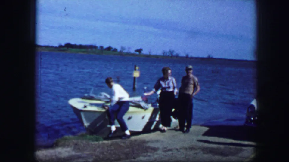 Final frame shows three adults beside the small boat at a lake dock, with a marker post, shoreline, and partial car visible.