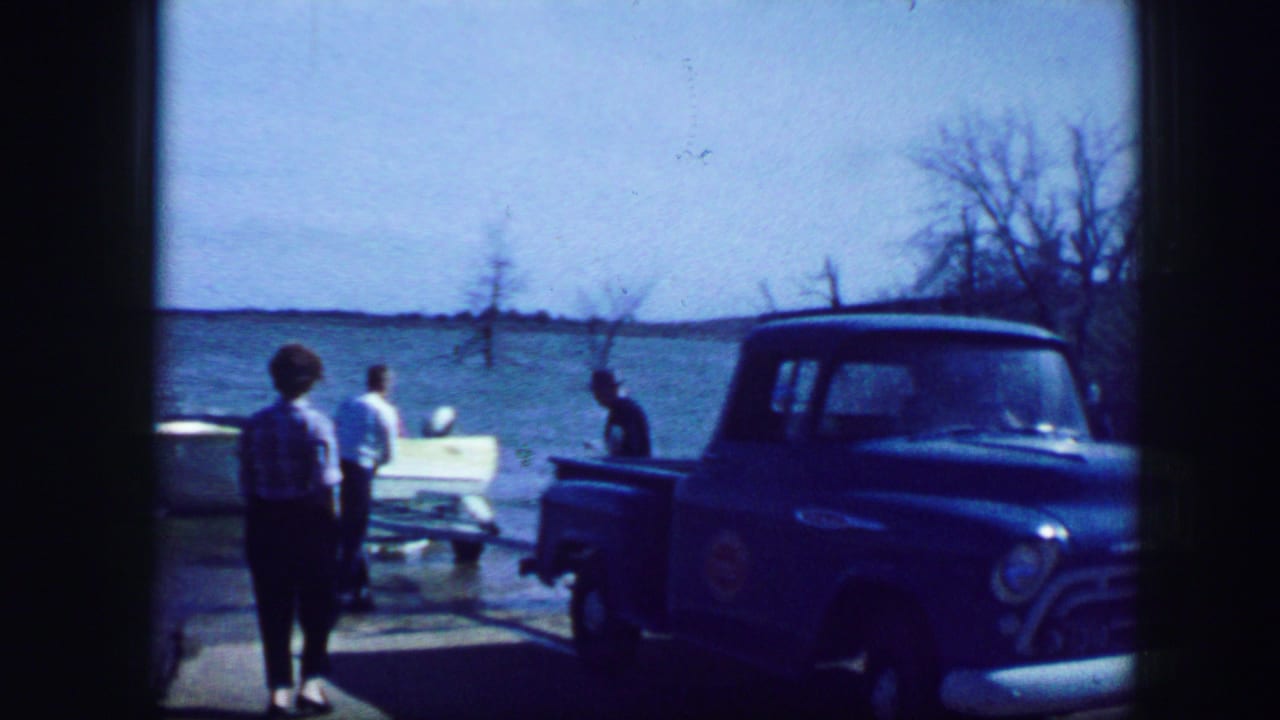 1964 Fort Worth home-movie frame with people near a white boat and blue pickup truck beside a bright lakeside ramp.