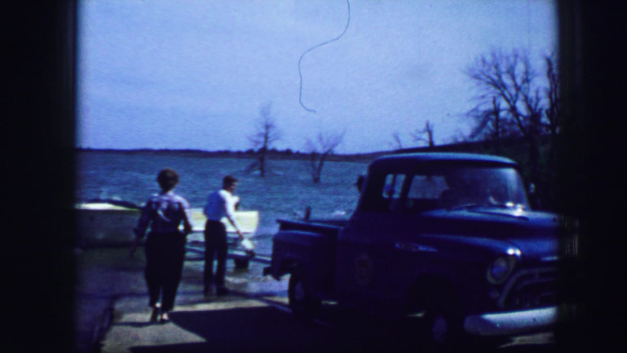 Two people approach a white boat beside a blue pickup truck near the water.