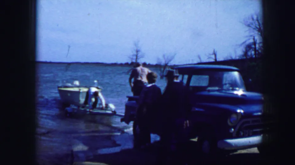 Home-movie frame from Fort Worth in 1964 showing people around a small white boat beside a blue pickup at the lake.