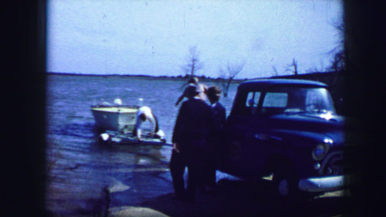 1964 Fort Worth-area home-movie frame showing adults near a blue pickup truck and small white boat at a lakeside.