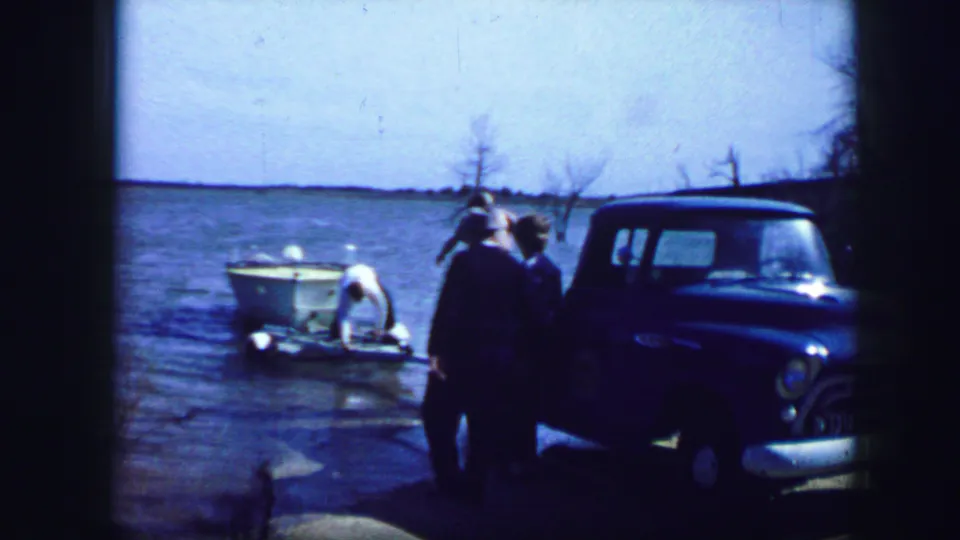 1964 Fort Worth-area home-movie frame showing adults near a blue pickup truck and small white boat at a lakeside.