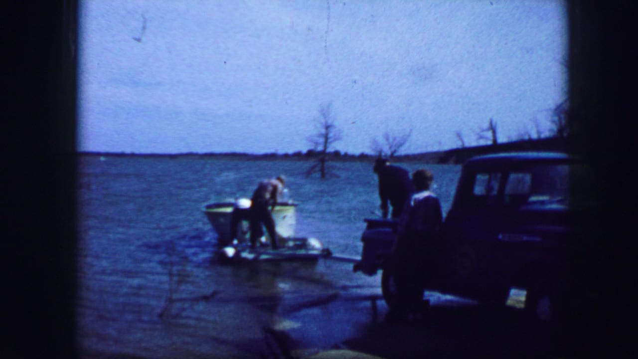 Person tending a small boat as others stand near a blue pickup at the lake.