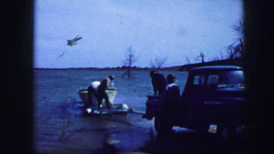 1964 lakeside home-movie frame with a small boat at the water's edge, adults nearby, and a blue pickup parked on shore.