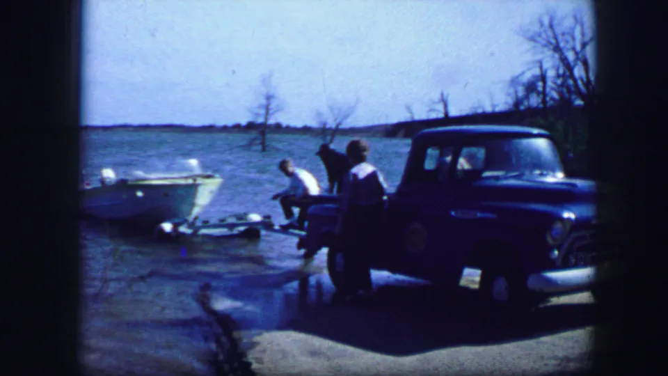 Fort Worth-area 1964 home-movie frame showing adults by a blue pickup and a person crouched or seated near the small boat at the lake.