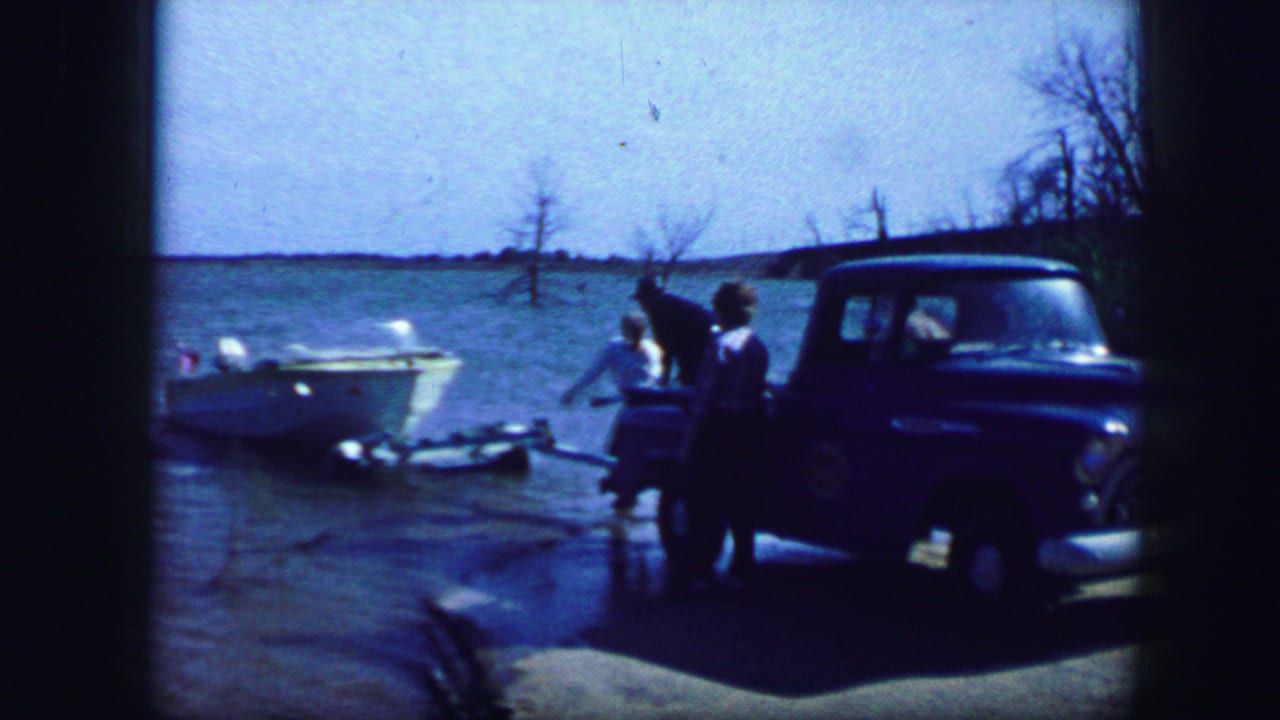 1964 Fort Worth home-movie frame showing a blue pickup at the shore with a white motorboat, open water, and people standing nearby.