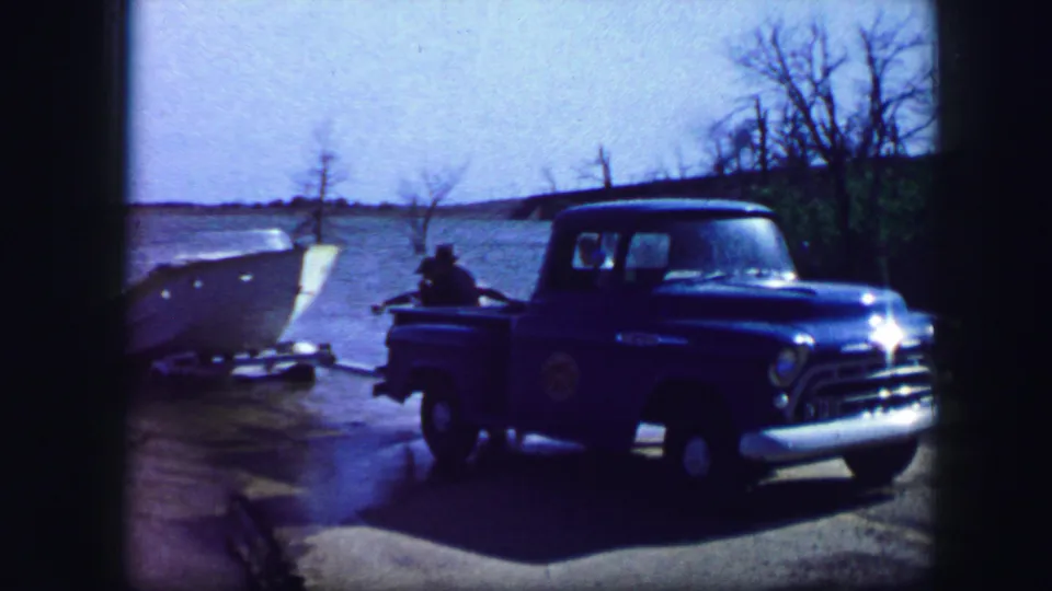 1964 Fort Worth home-movie frame showing the front of a blue pickup near a white motorboat and lakeside shore.
