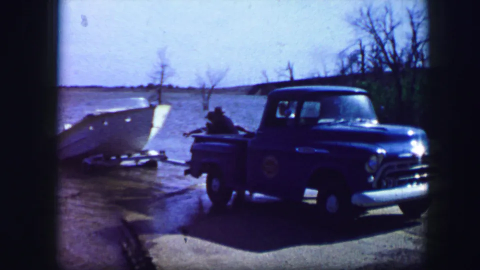 1964 Fort Worth frame showing a blue pickup at the shoreline, a person in the truck bed, and a white motorboat beside open water.