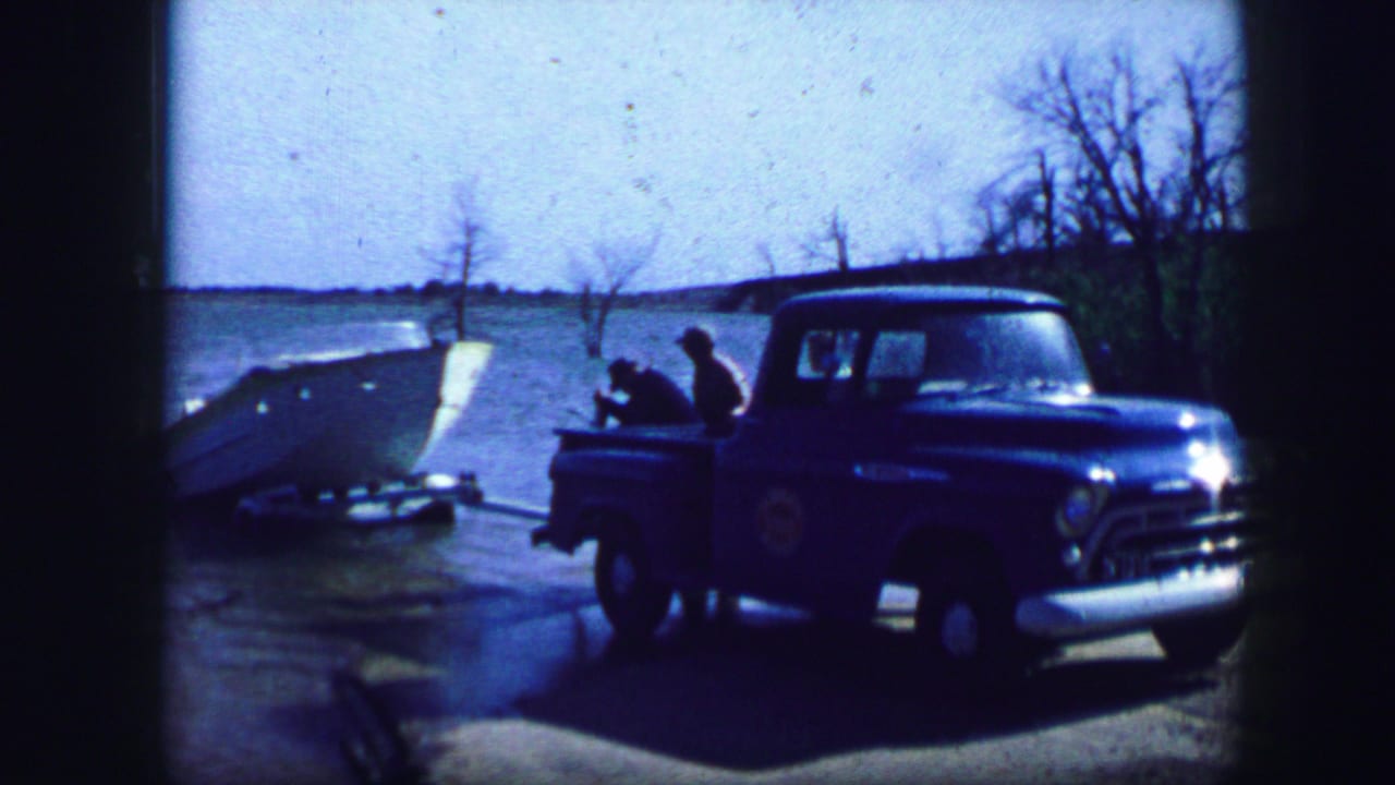 Blue pickup truck towing a small boat beside a body of water.