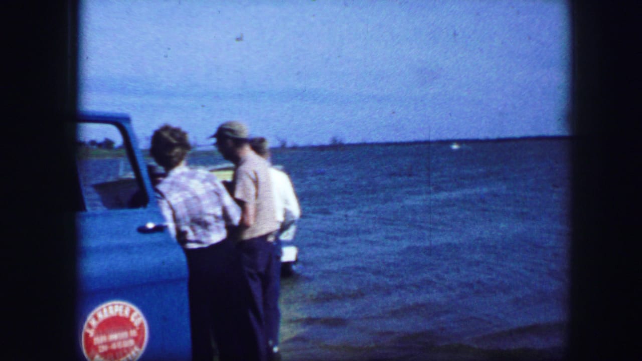 Adults stand beside the open door of a blue truck near the water.