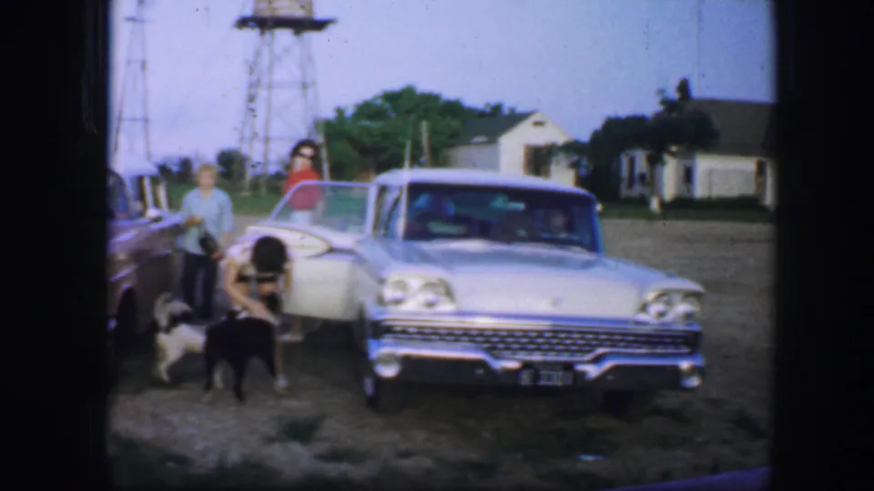 1964 Fort Worth-area home-movie frame showing two people with dogs beside a light-colored vintage car near small buildings.