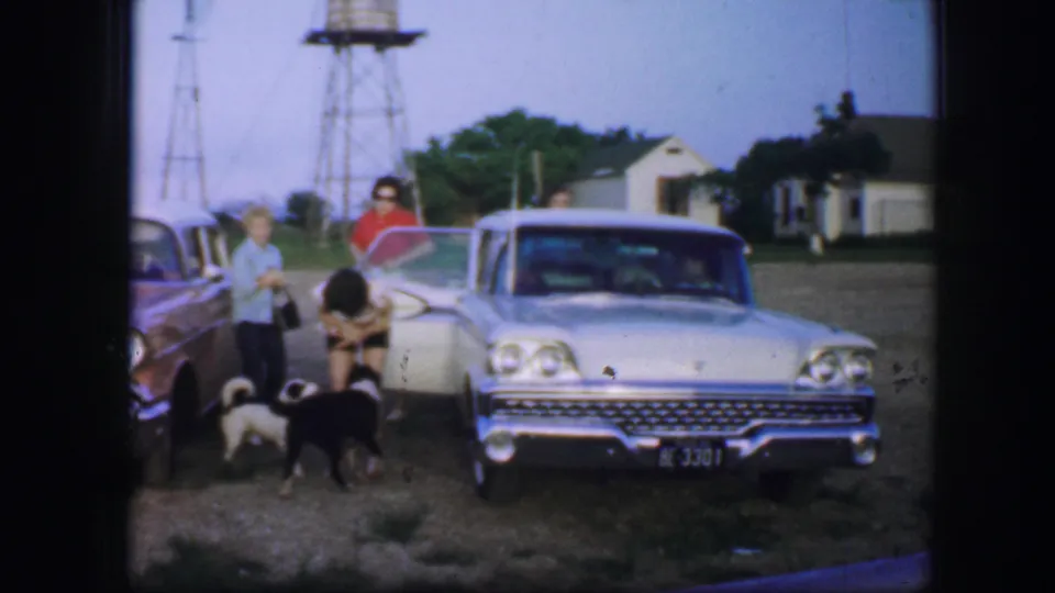 1964 Fort Worth-area frame with people and dogs in front of a vintage car, set near water towers and small roadside buildings.