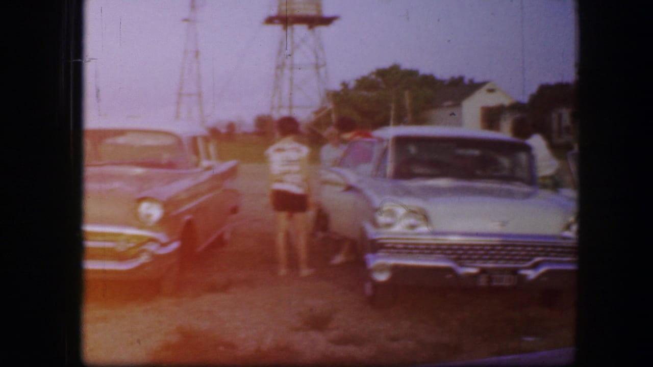 Two cars parked side by side as people stand near the vehicles under a bright sky.