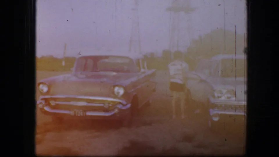 Fort Worth 1964 home-movie frame with a front-facing car, another car beside it, a standing person, and sun-faded 8mm exposure.
