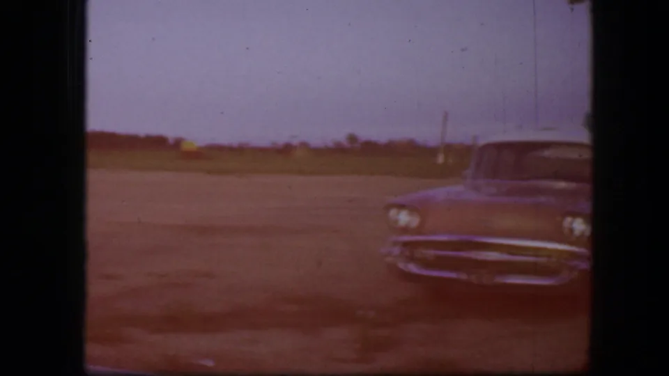 1964 Fort Worth home-movie frame showing one parked car at the right edge of a wide open lot beneath a pale sky.