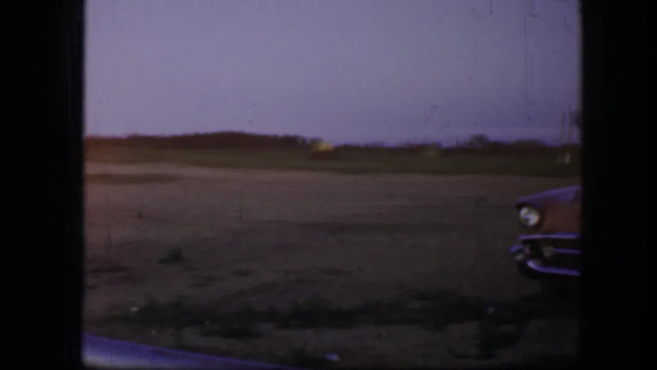1964 Fort Worth home-movie frame showing an open field at dusk, distant horizon, dark film edges, and part of a car front at the right edge.