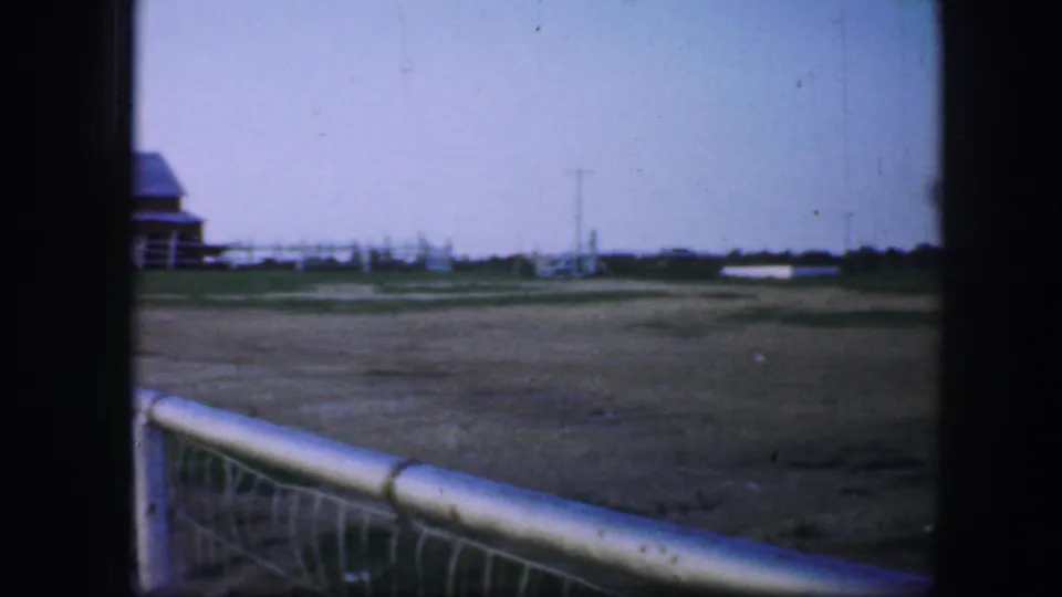 1964 Fort Worth home-movie frame showing a broad field, distant barn-like building, fence line, utility pole, and pale foreground railing.