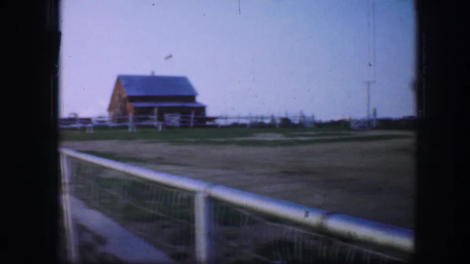 1964 Fort Worth frame showing a barn-like building beyond open ground, with fence rails, a utility pole, and a pale railing in the foreground.