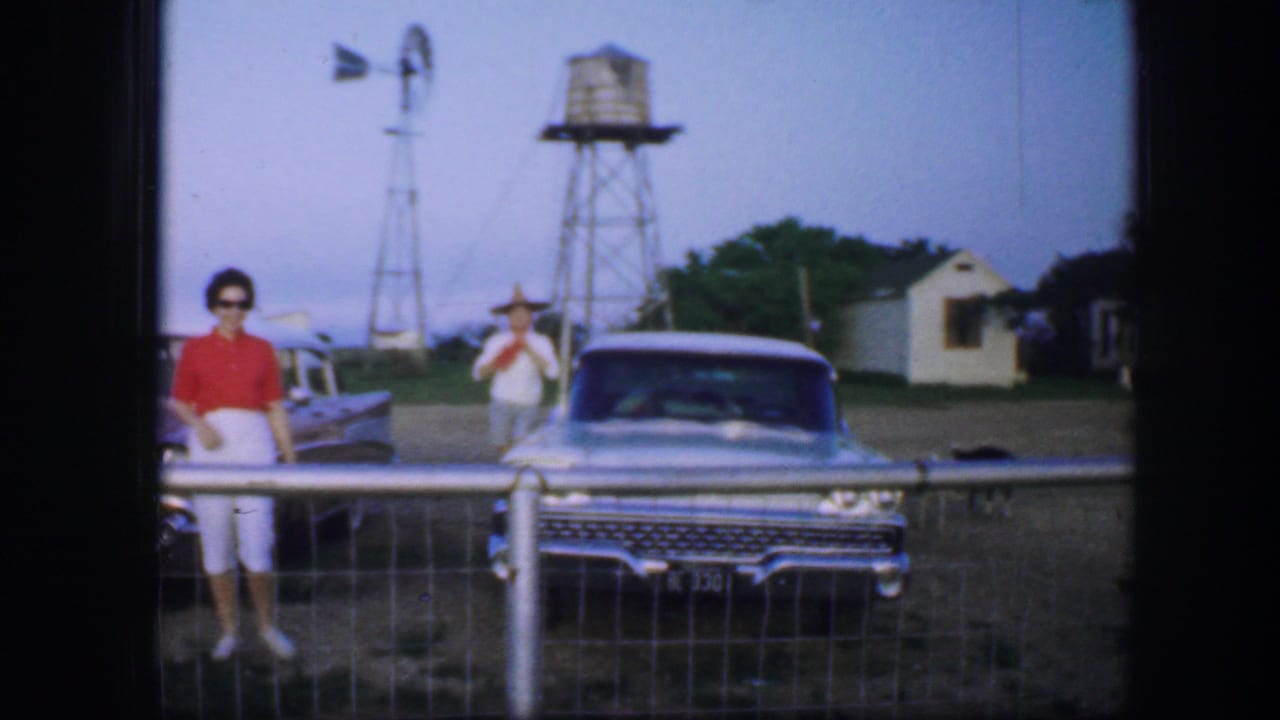 Two women stand near classic cars behind a farm fence with a windmill and water tower in the background.