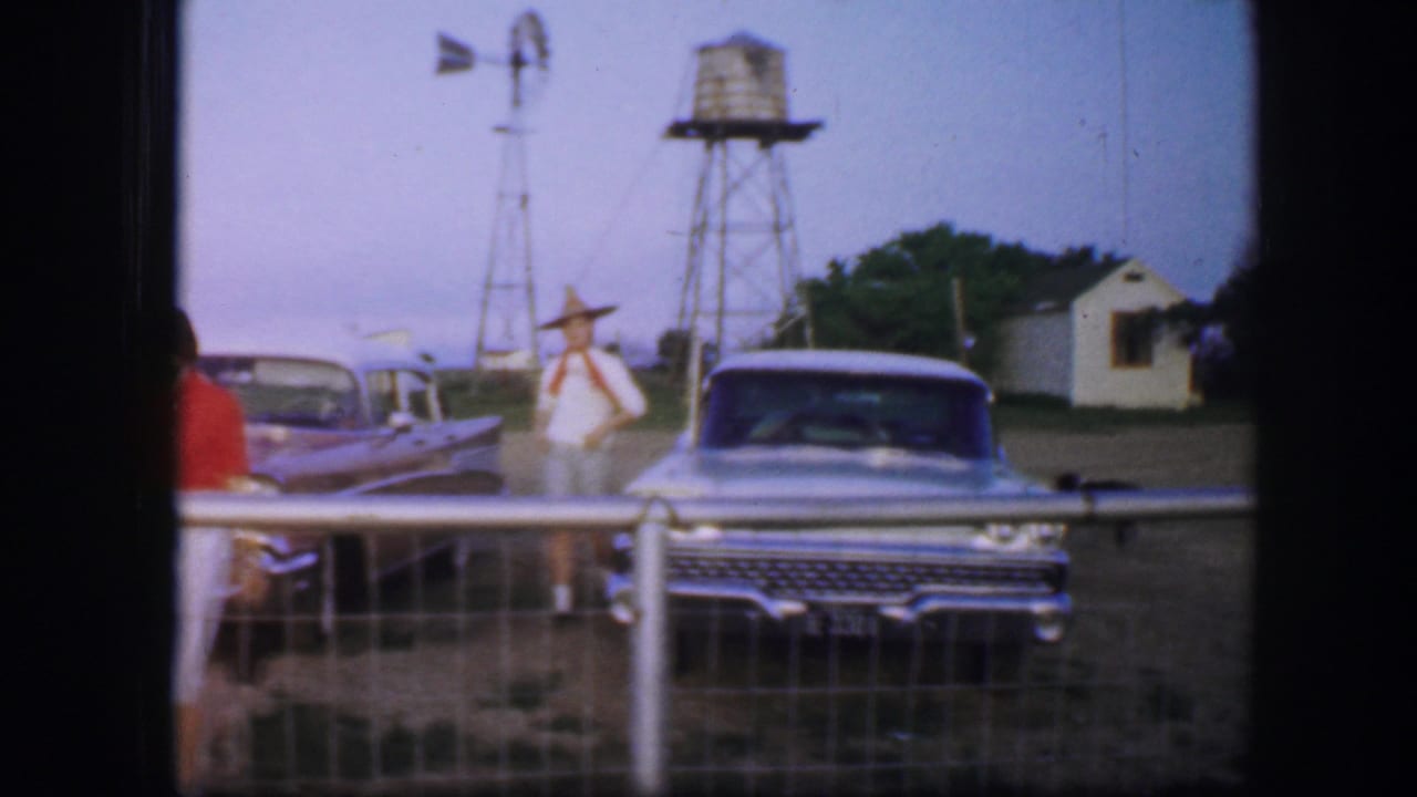 A woman in red stands partly turned away while another woman in a hat stands near parked cars and farm structures.