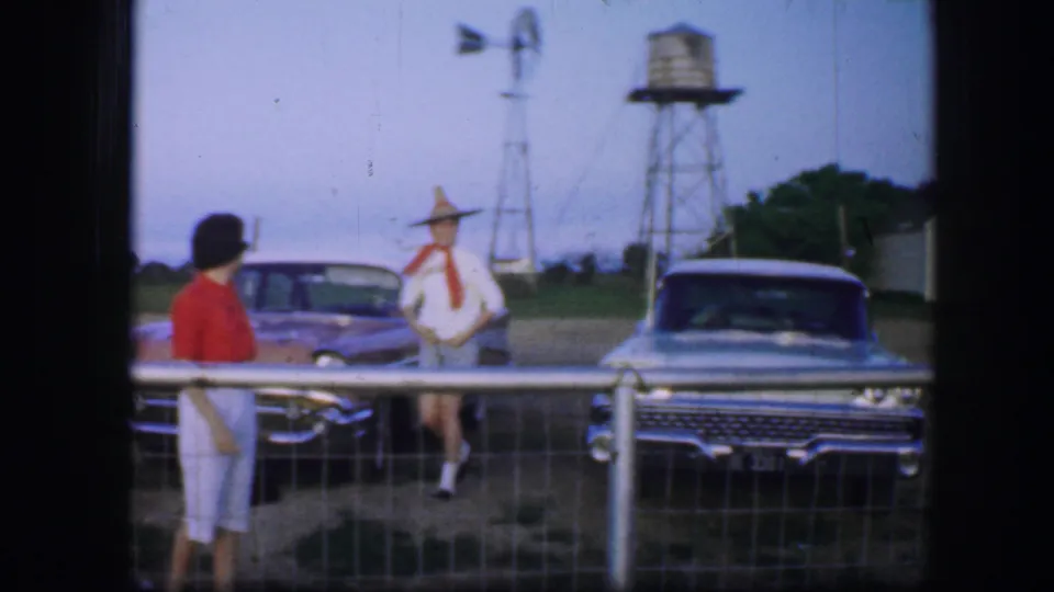1964 Fort Worth-area home-movie frame of two adult women near classic cars, with a fence, windmill, water tower, and rural buildings behind them.