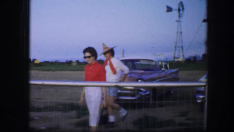 1964 Fort Worth-area frame showing two women walking near a fence and parked classic car, with a windmill and rural landscape behind them.