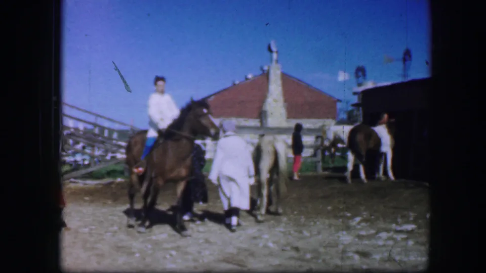 1964 Fort Worth home-movie view of a mounted rider, handlers in light coats, and horses gathered in a dusty fairground area.