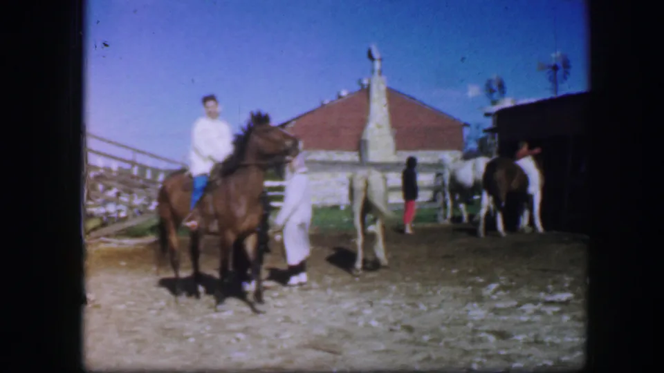 1964 Fort Worth frame showing a rider on a brown horse, a handler, and several horses near a red-roofed barn and fence.