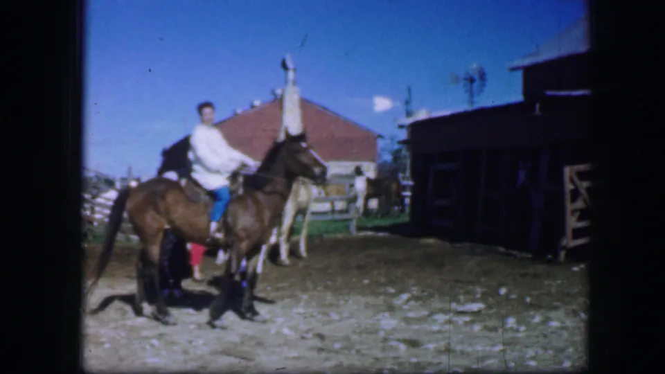 1964 Fort Worth footage frame with a rider on horseback near a light horse, two people, fencing, and stable-like buildings.