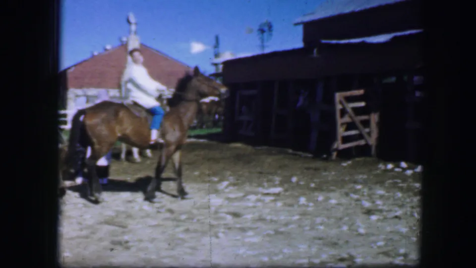 1964 Fort Worth home-movie frame showing a rider on a dark horse near a shaded stable building and fenced ranch yard.