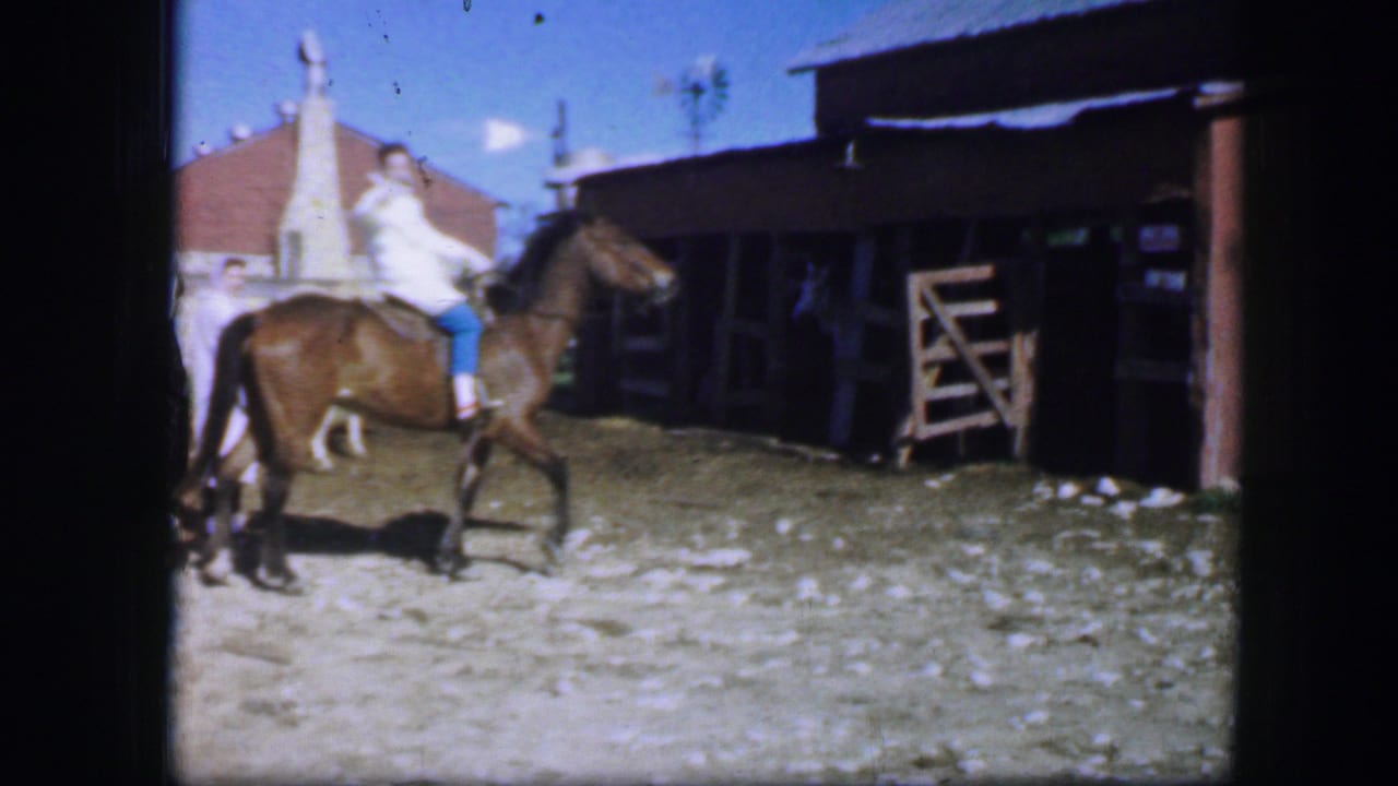 In Fort Worth, Texas in 1964, a woman in a white coat rides a brown horse past a wooden stable area on dusty ground.