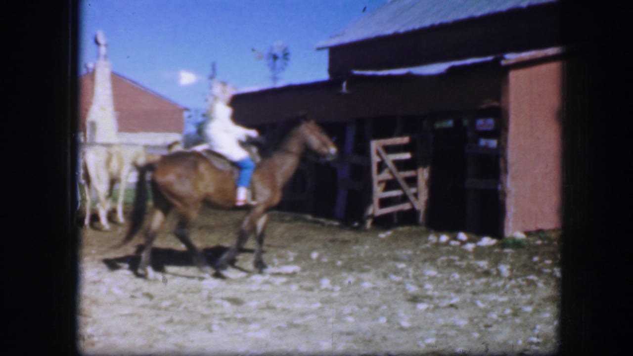 Woman waves while riding a brown horse past a wooden structure.