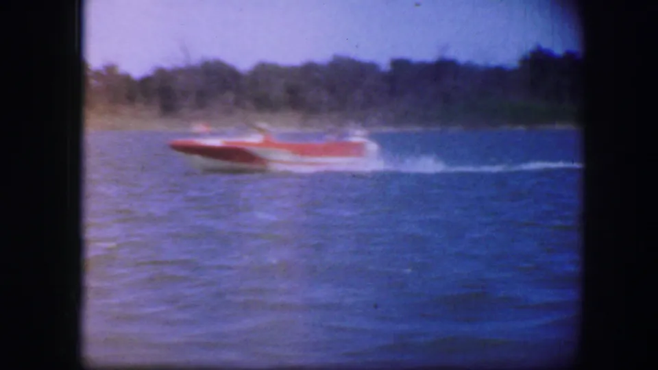 A red speedboat crosses open water near a wooded shoreline in a soft 8mm home-movie frame associated with the same clip.