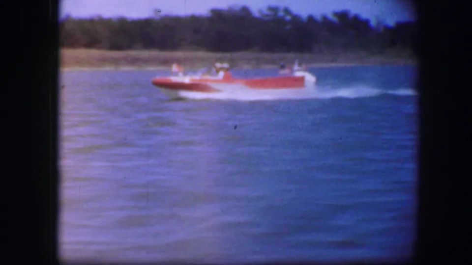 A red speedboat carrying several people moves across the water near a tree-lined shore in the 1960s home-movie footage.