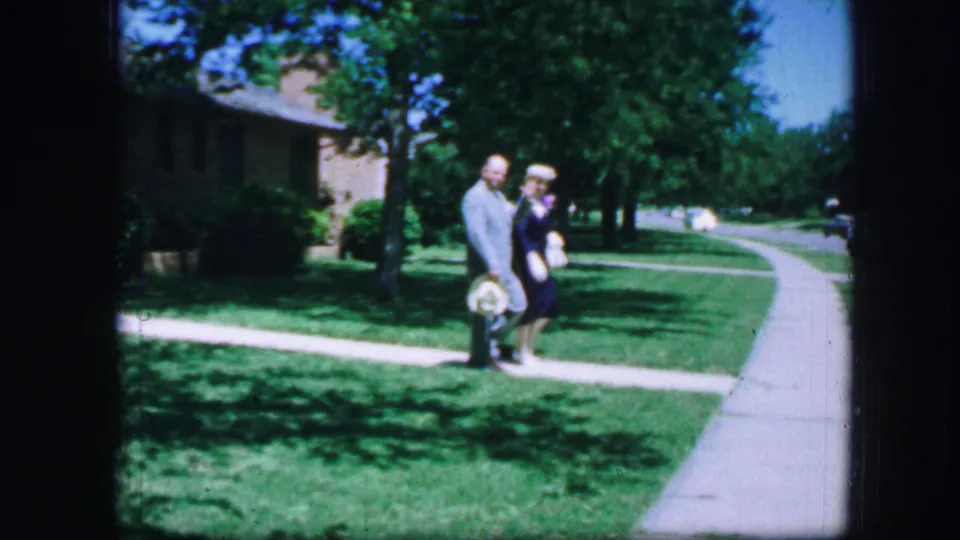 An adult couple in mid-century dress stands or slows near a sidewalk crossing, with grass, trees, and a residential street beyond.