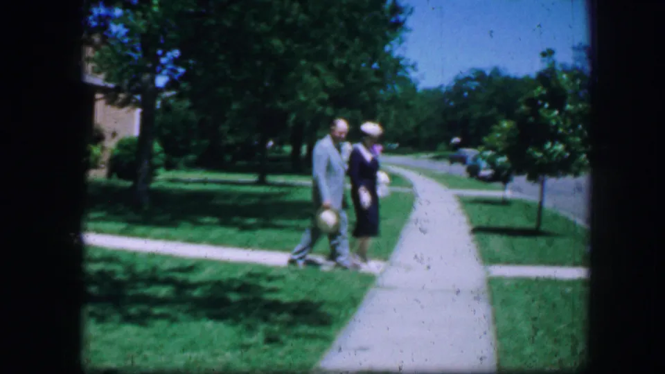 The couple walks past a curving suburban sidewalk in Fort Worth, framed by lawns, trees, and a quiet street in the background.