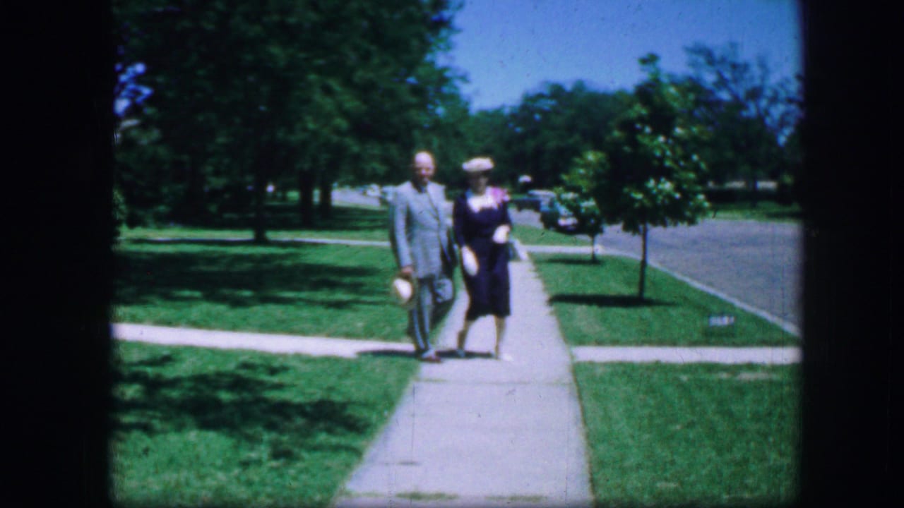 In 1964 Fort Worth, an adult couple in formal clothing walks along a residential sidewalk bordered by lawns and trees.