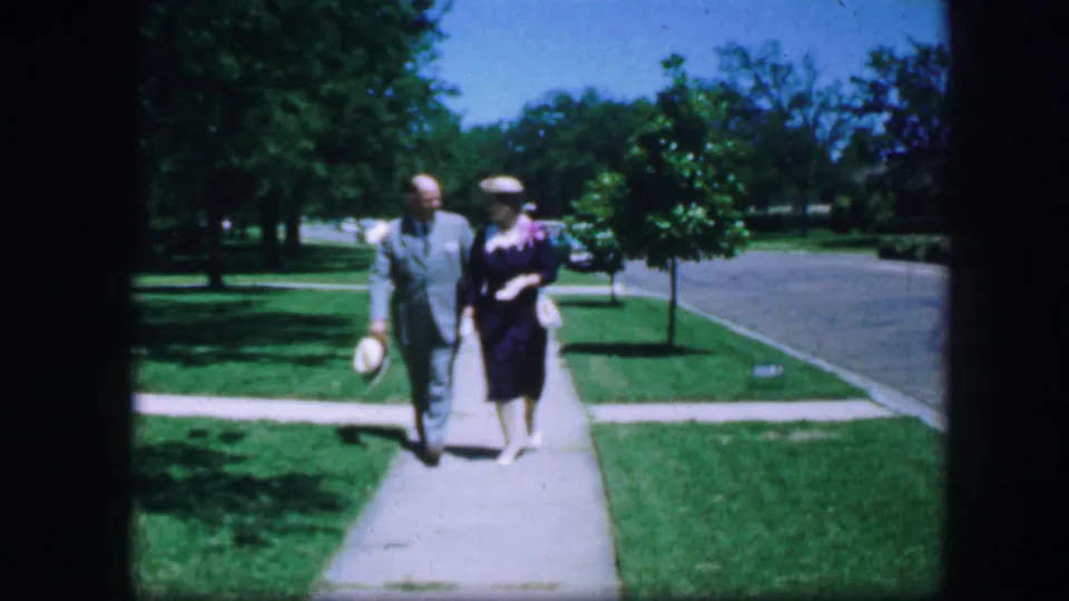 The man in a light suit and the woman in a dark dress and hat continue toward the camera in a Fort Worth residential setting.