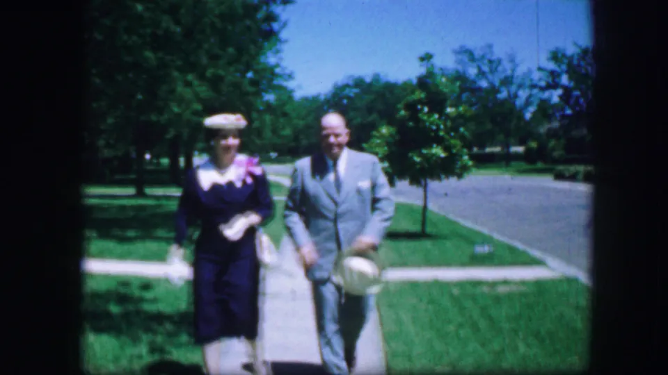 Near the end of the shot, the adult couple passes close to the camera, the man carrying a hat and the woman wearing a dark dress and hat.