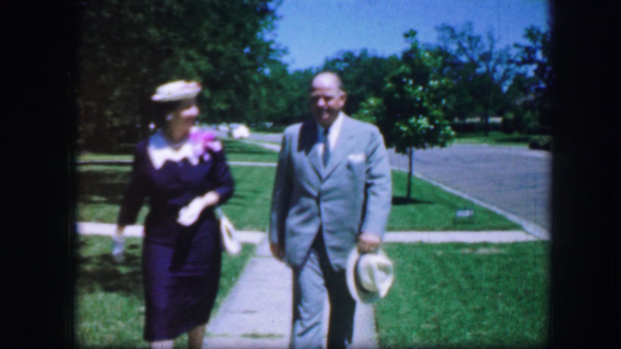 In 1964 Fort Worth home-movie footage, a woman in a blue dress and hat walks beside a man in a gray suit holding a white hat.