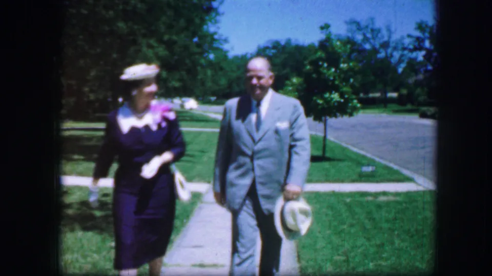 In 1964 Fort Worth home-movie footage, a woman in a blue dress and hat walks beside a man in a gray suit holding a white hat.