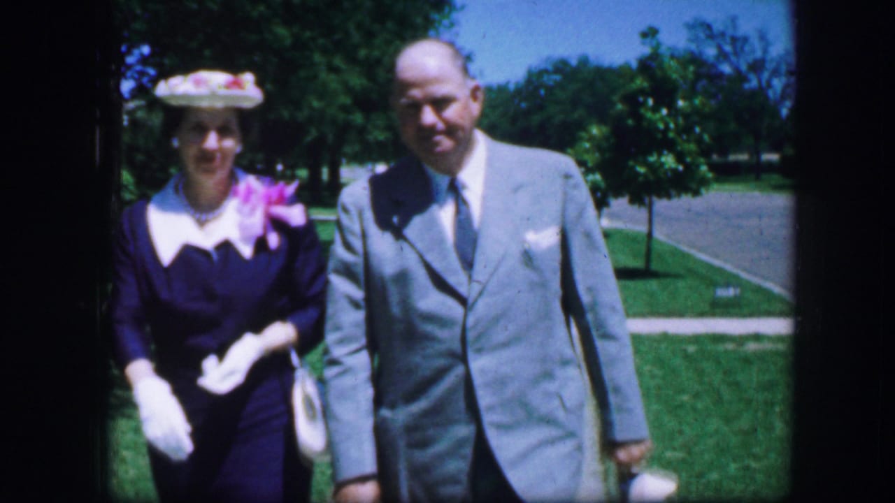 Couple in formal clothing near the camera on a sidewalk with lawn and trees behind them.
