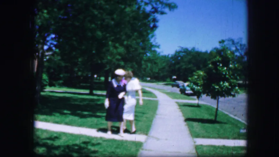 Two women, one in a blue dress and one in a light outfit, pause together on a sunny Fort Worth sidewalk in 1964 home-movie footage.