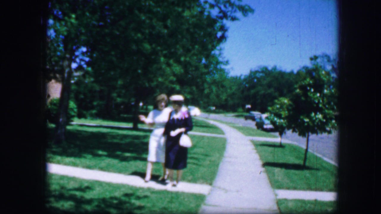 Two women stand on a suburban sidewalk beside grass and trees.