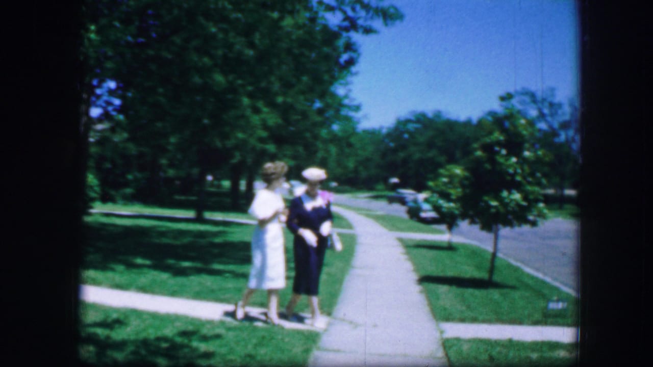 Two women face each other on a sidewalk near a quiet street.