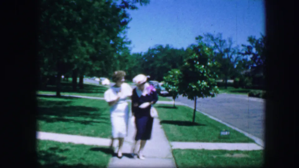 Two women continue walking together on a Fort Worth residential sidewalk, framed by green lawns, trees, and a quiet street.