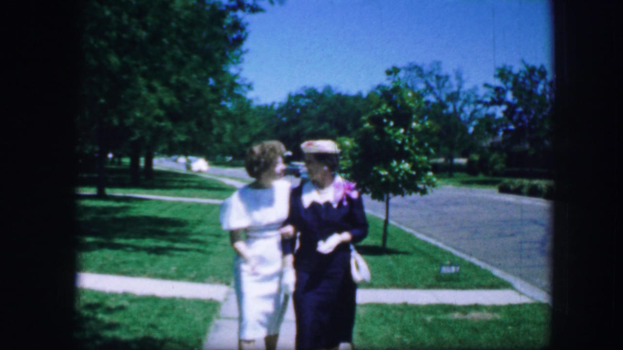 Two women walk arm in arm beside a sidewalk, one wearing a floral hat and gloves.